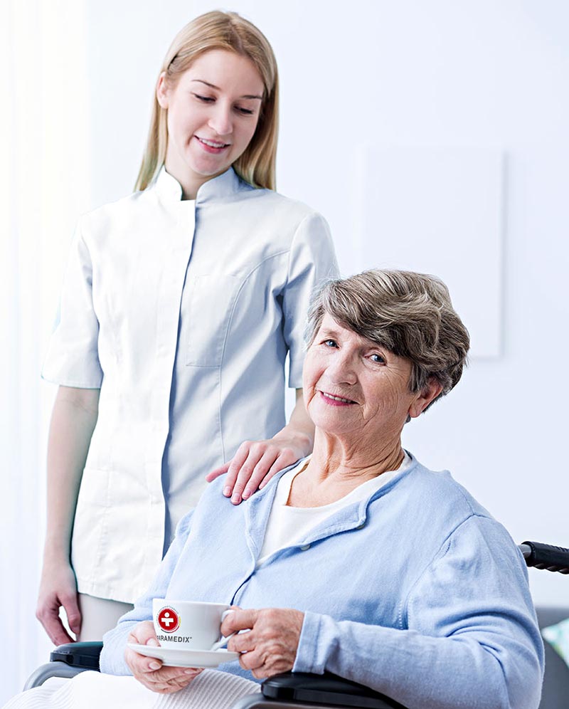 Eine Pflegehelferin steht neben einer Patientin im Rollstuhl, die eine Tasse mit dem Paramedix-Logo hält.